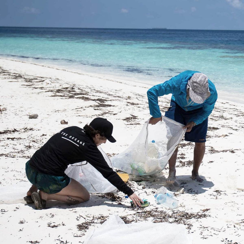 Two people holding rubbish bags are collecting plastic bottles off a beach shore. 