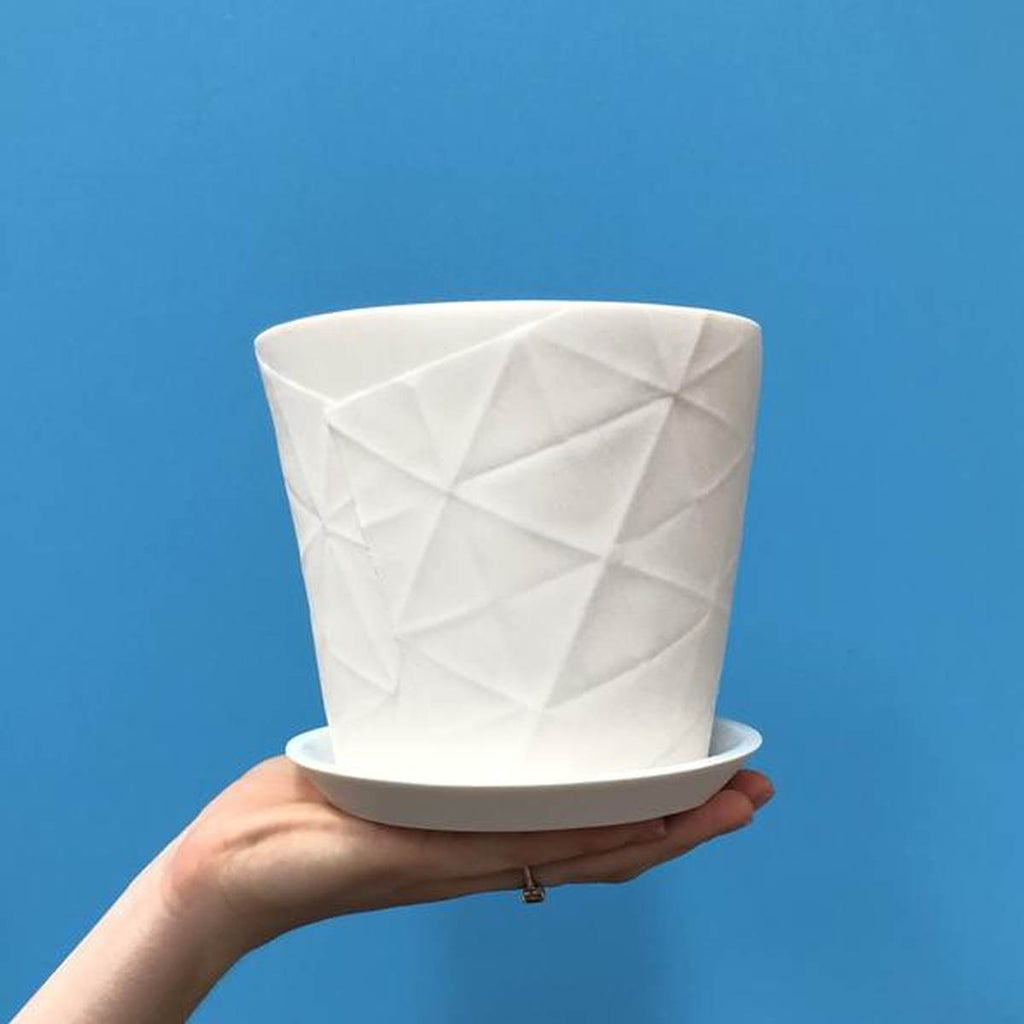 White porcelain planter with the imprinted creases from an unfolded chatterbox paper and a white dish held up by a hand against a blue wall. 