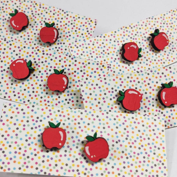 A pair of intricately hand coloured studs depicting rosy red apples with green leaves. The peeled layers hang down. Displayed on a rainbow polka dot background.