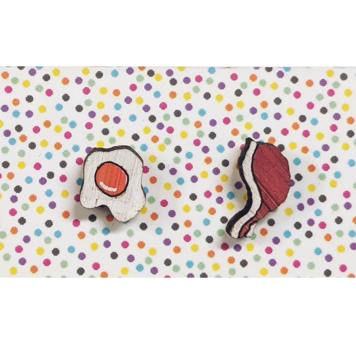 A pair of intricately hand coloured studs one in the form of a fried egg, the other a rasher of bacon. Shown on a rainbow polka dot background.
