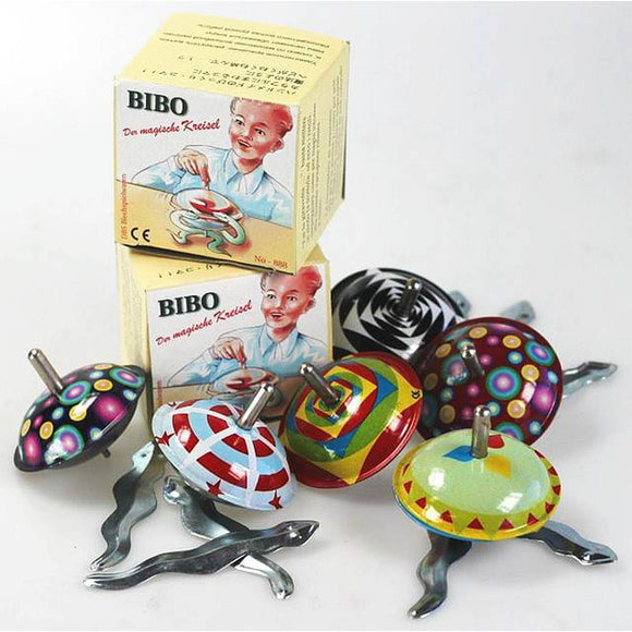 Three brightly coloued tin spinning tops, pictured in front of a pile of boxes. The boxes feature the text " Bibo Der Mahische Kreisel" and an illustration of a child playing with a spinning top. There a metal snake shape pieces scattered around the spinning tops.