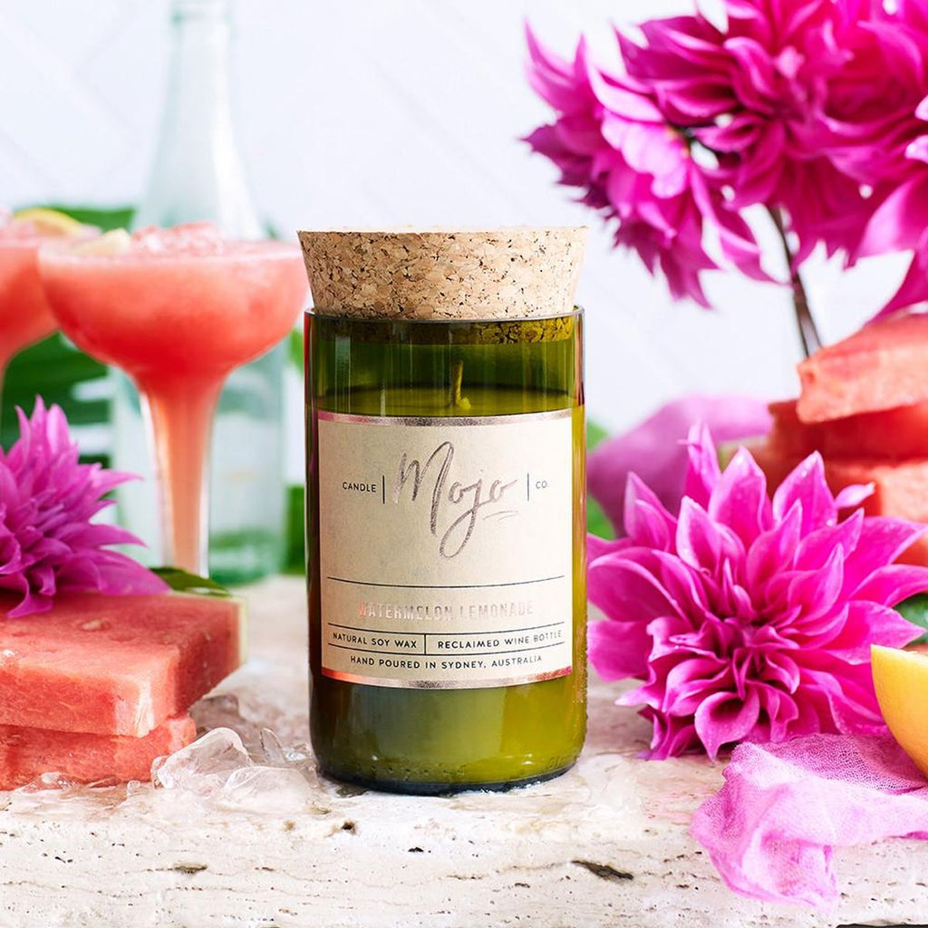 The bottom half of a recycled green wine bottle with a cork top contains the soy candle stands in the centre, framed with a glass of watermelon cocktail, watermelon slices, ice blocks and pink flowers. 